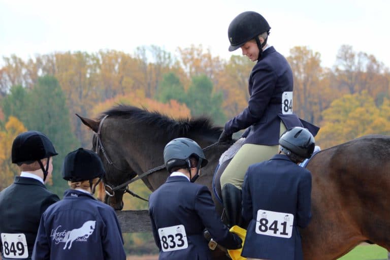 A rider mounted on a horse while teammates polish their boots